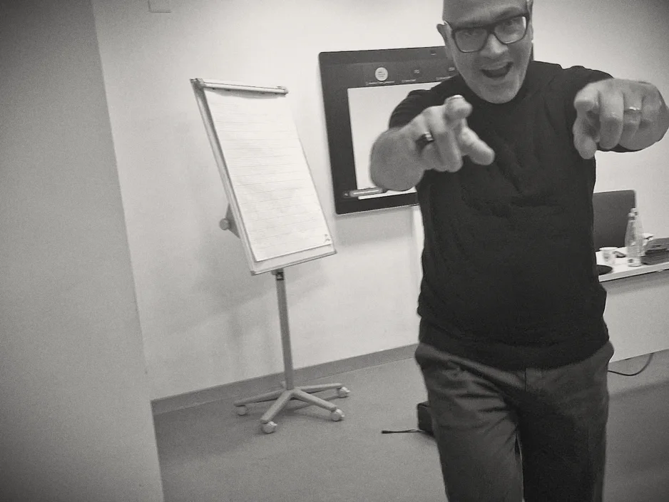 Gabriele Micozzi in aula indica l'obiettivo con tutte e due le mani, davanti a una lavagna a fogli mobili